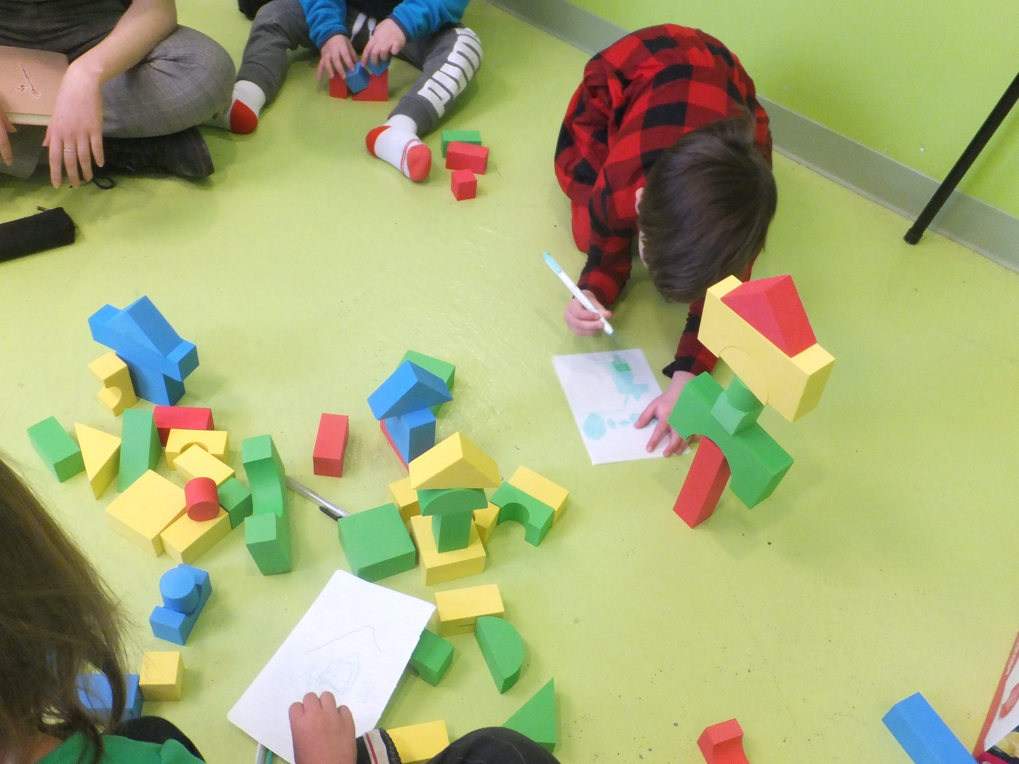 Trois enfants et une adulte sont assis autour des blocs de constructions, des feuilles de coloriages, et des marqueurs.